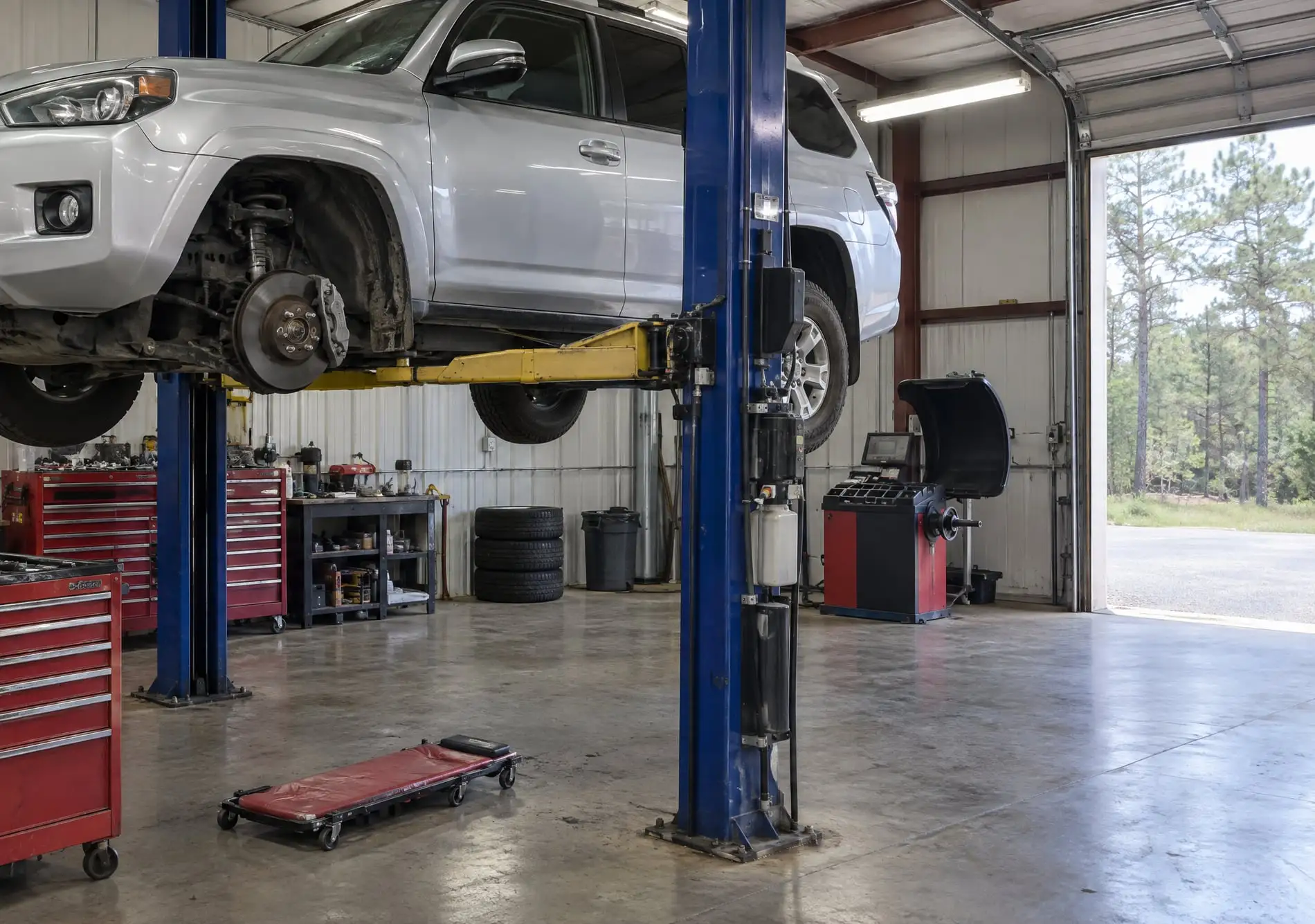 Inside a shop bay: an SUV on a two-post lift with a front wheel off, red tool chests, a tire balancer, the bay door open to the pines