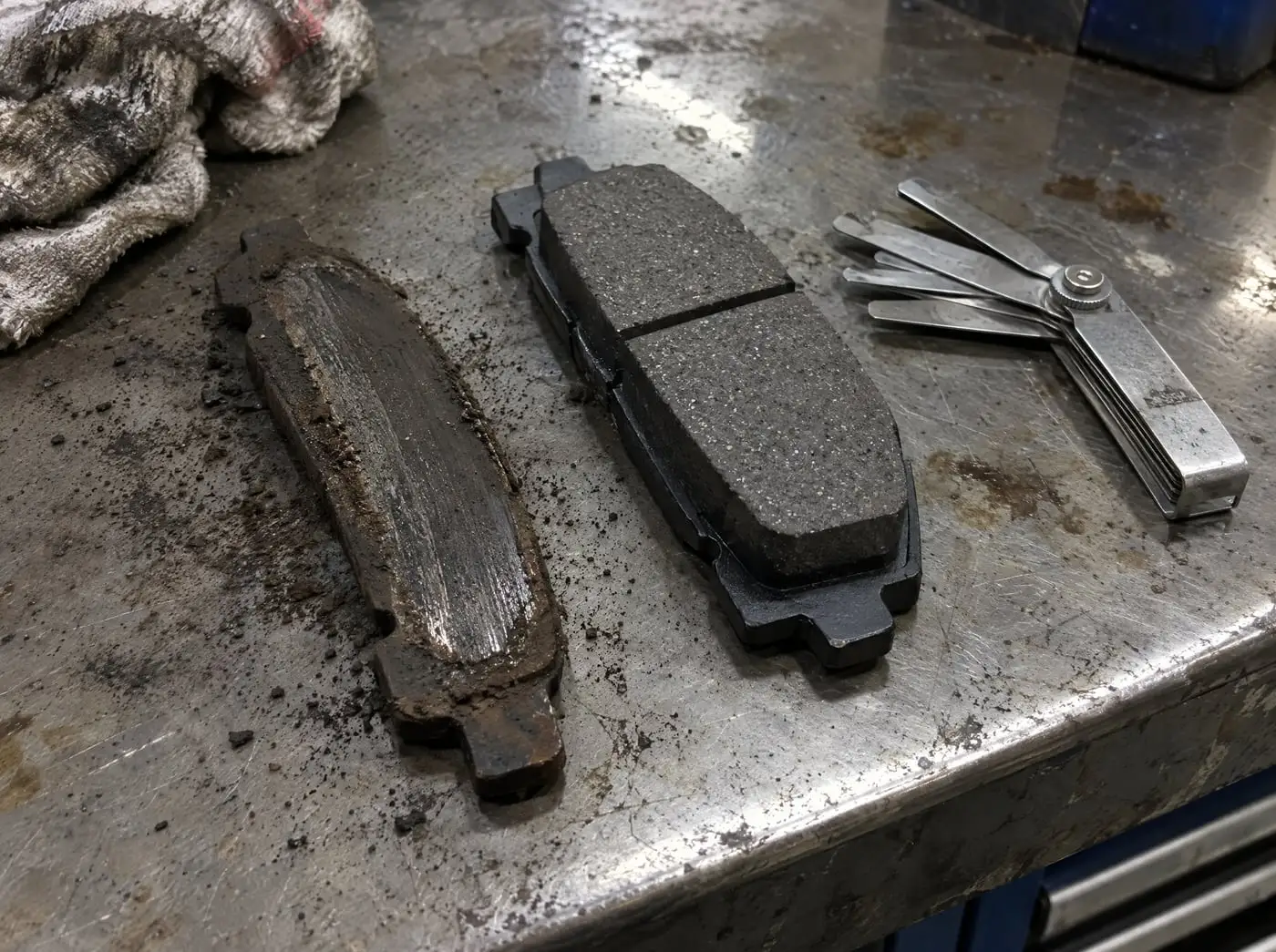 A worn brake pad worn to a thin sliver beside a new pad on a workbench, with a thickness gauge