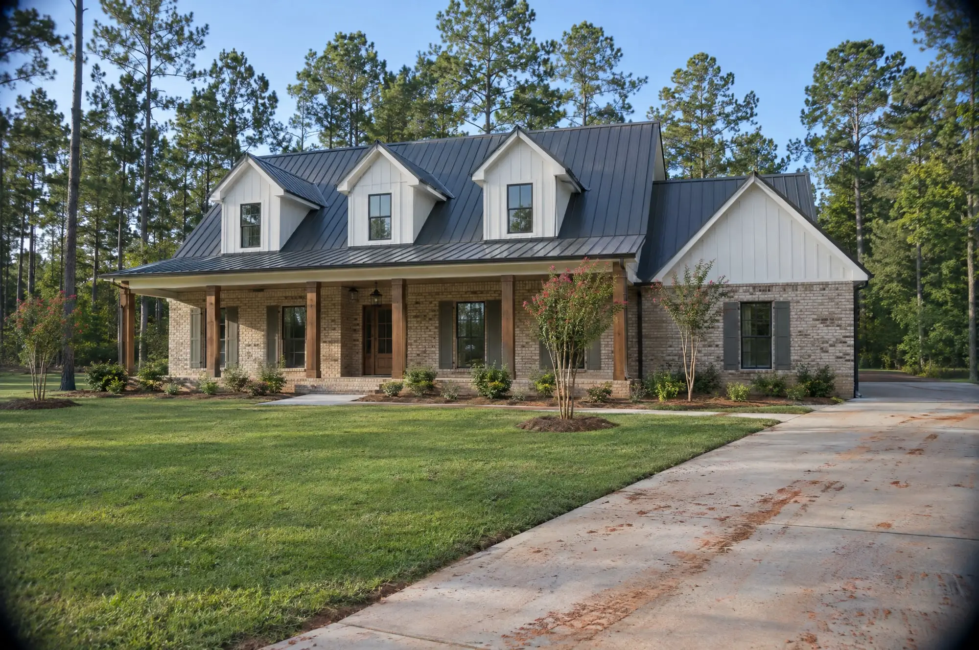 A brick story-and-a-half house with three dormers in a dark metal roof and a long front porch, red clay tracked onto the drive