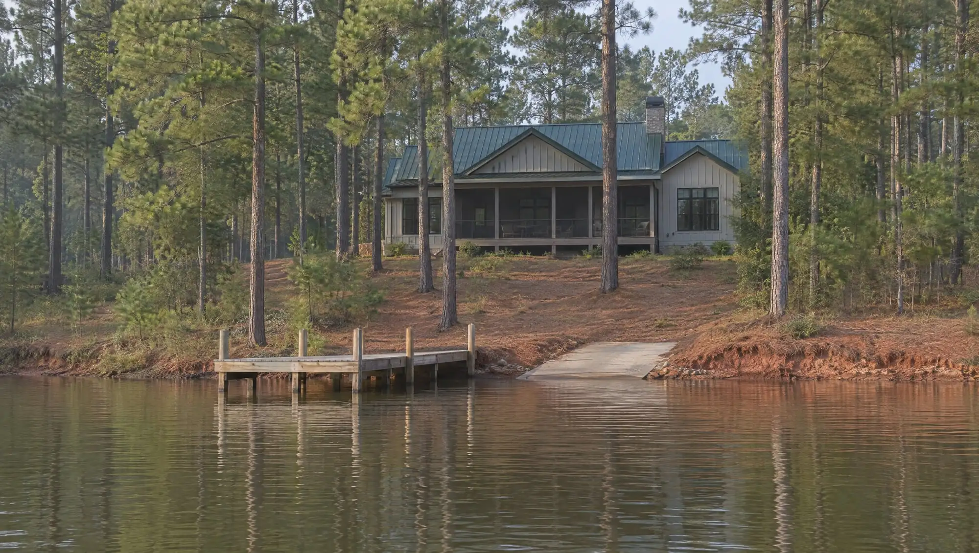 A lake house with a green metal roof and a screened porch up the slope in the pines, a wooden dock and a concrete boat ramp at the shore