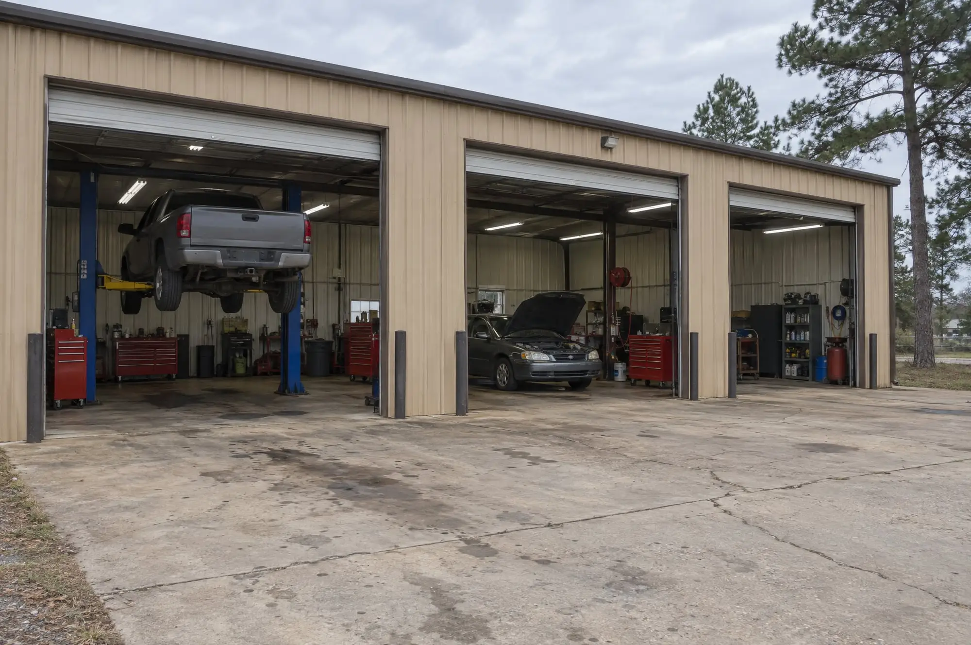 A tan metal shop with three bay doors up: a pickup on a lift, a sedan with its hood open, red tool chests inside