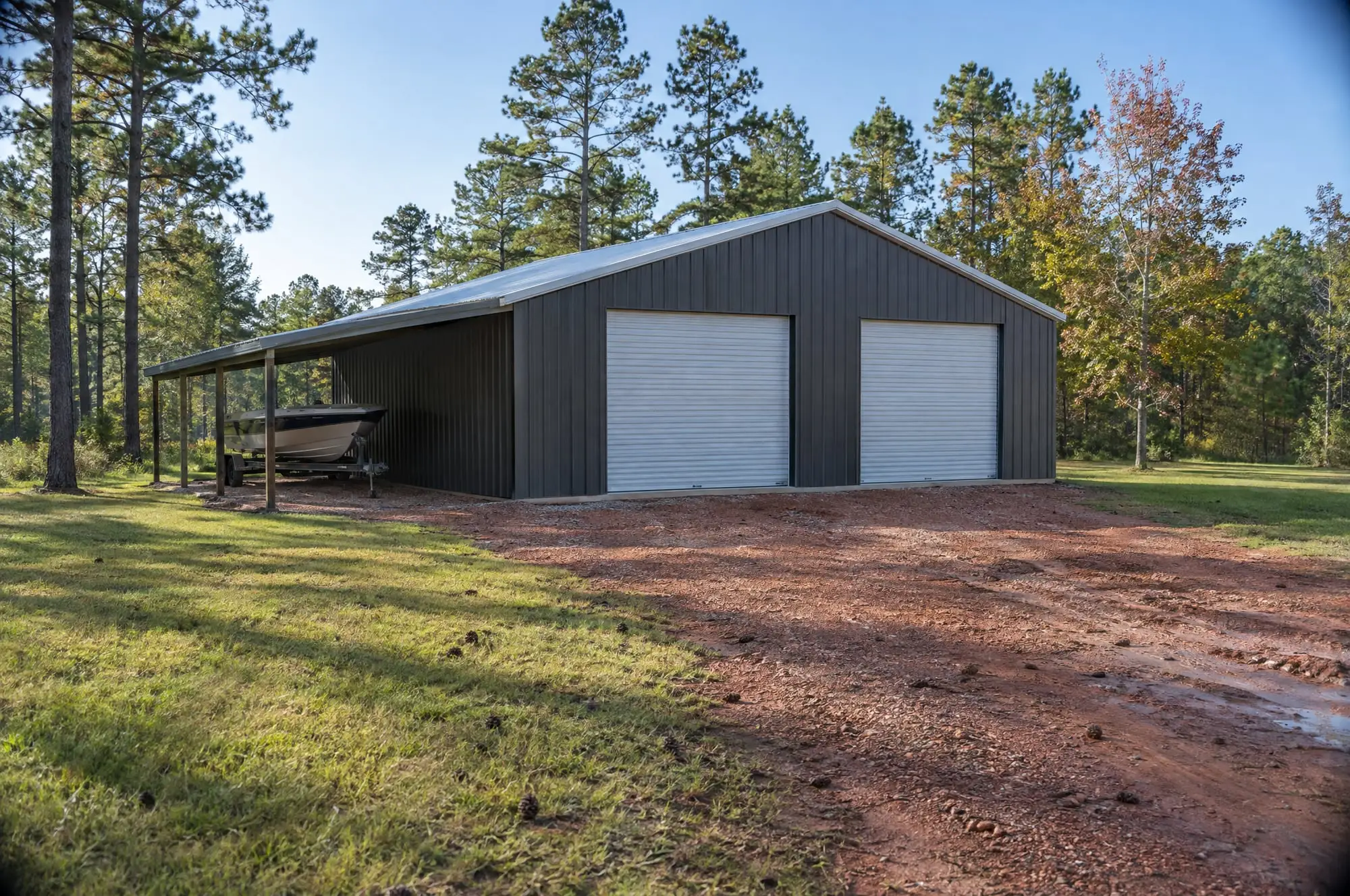 A charcoal steel shop with two white roll-up doors and a lean-to sheltering a boat, on a red clay drive in the pines
