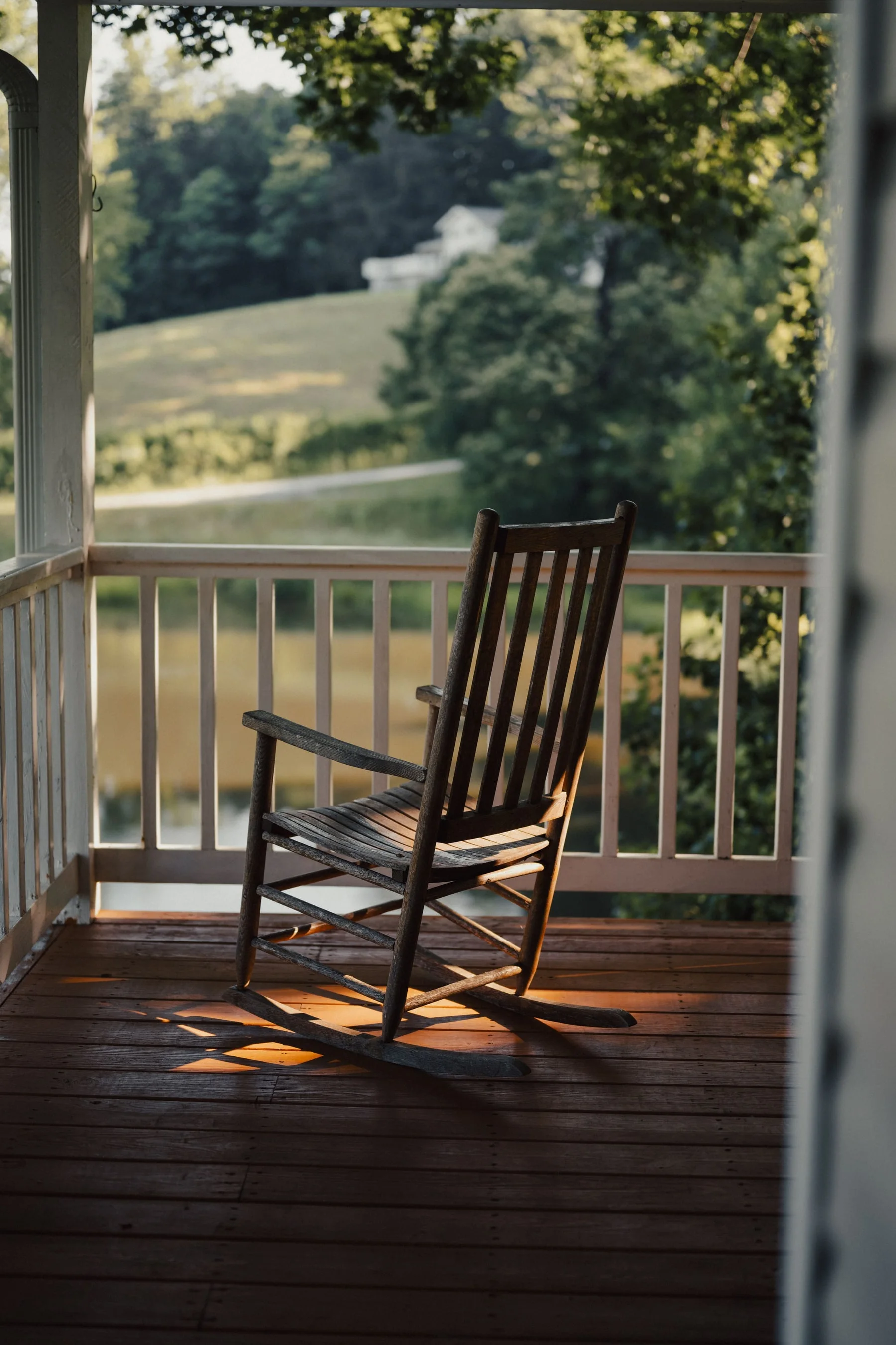 An empty wooden rocking chair on a porch in late sun, looking out over a pond and green hills