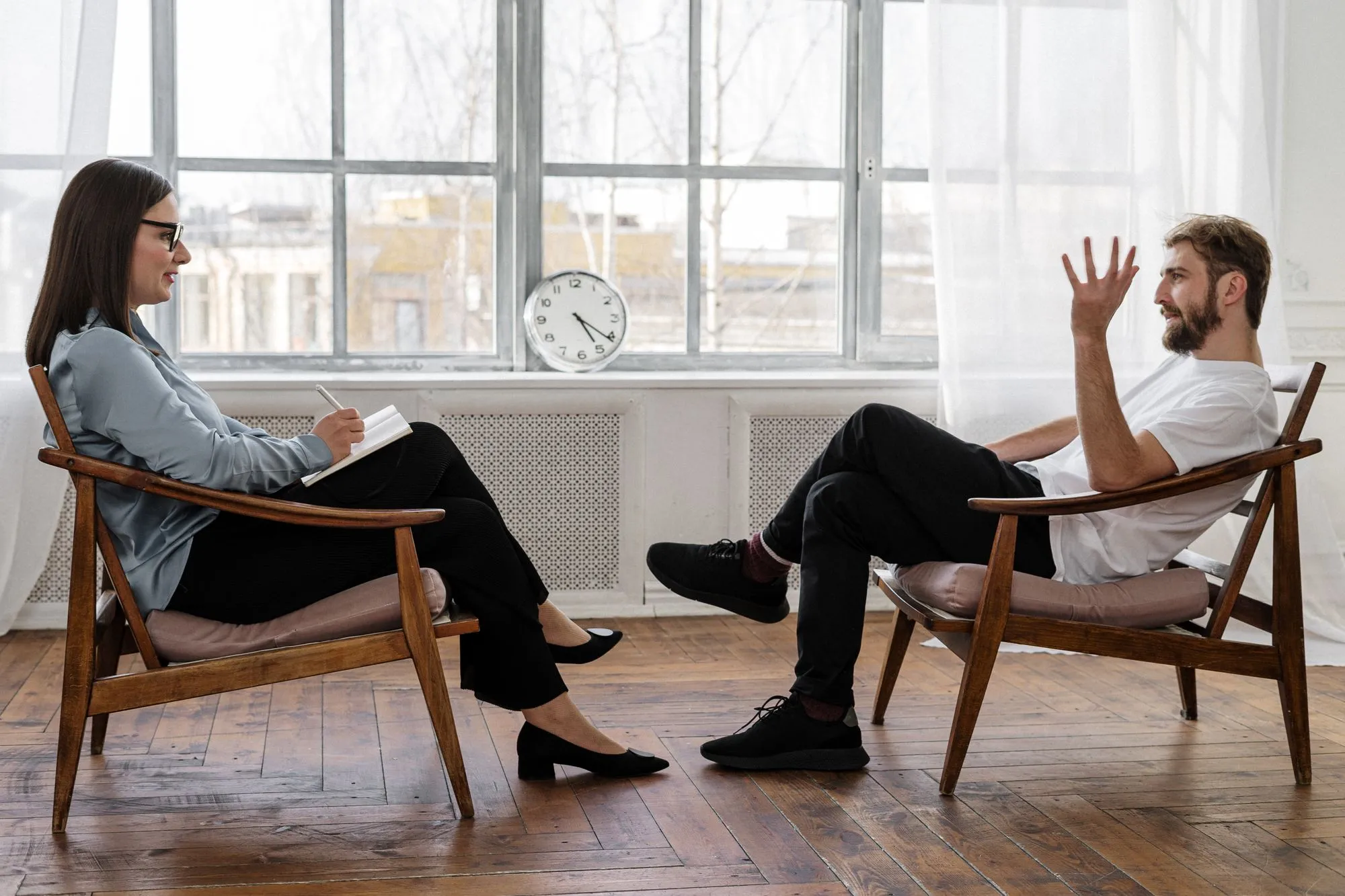 A man in a bright room with tall windows, talking with his hands while a counselor listens