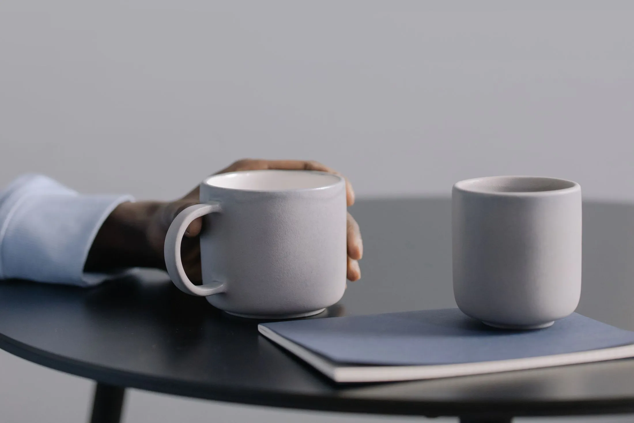 Two coffee mugs on a small table beside a notebook, one held by a hand
